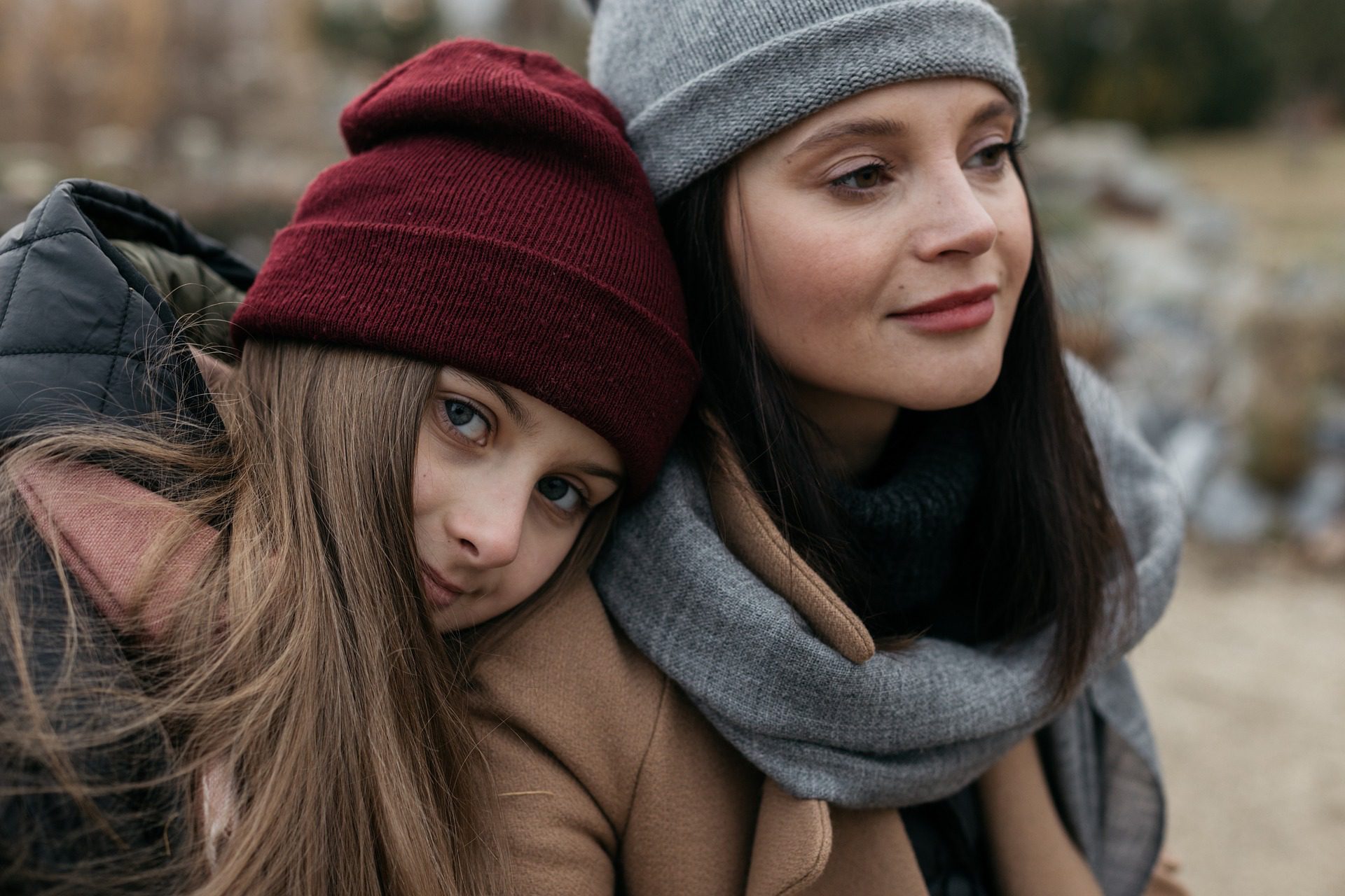 Two girls wearing hats and scarves leaning on one another.